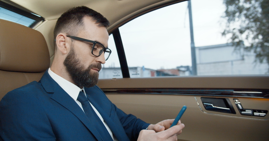 Young handsome Caucasian man in suit, tie and glases sitting on backseat of expensive car and holding cellphone. Serious businessman with beard and mobile phone, going somewhere and looking at window.