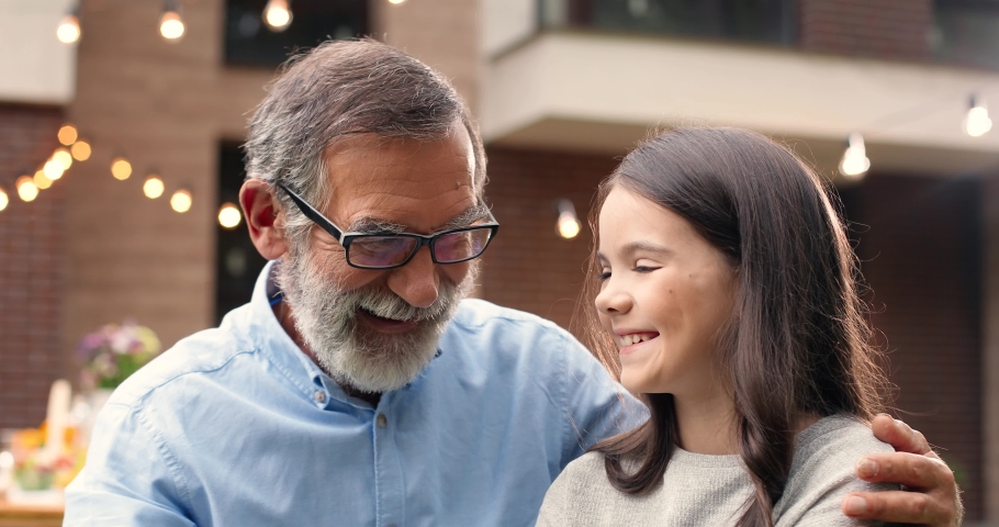 Portrait of Caucasian grandfather in glasses hugging small cute granddaughter and smiling in yard. Old gray-haired man embracing little pretty girl. Celebration outdoors. Family dinner and meeting. Royalty-Free Stock Footage #1064176954