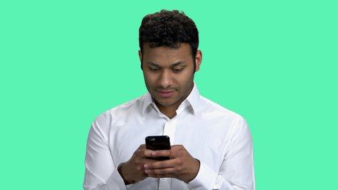 Smiling businessman typing message on his phone. Young Indian entrepreneur texting a message on his cellphone standing against color background.