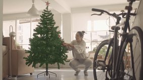 Young dark hair caucasian woman decorating a Christmas tree with red shiny balls. She calls a daschund puppy that runs in and out of the frame. Full still shot 4k high quality video footage.