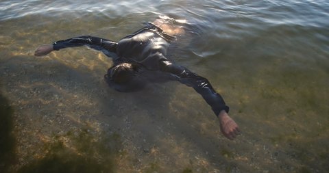 Drowned man with long dark hair in the water sways in the muddy water lying face down in shallow water. An unrecognizable body in black clothes is washed ashore after a shipwreck. 50 fps 4k slow motion.