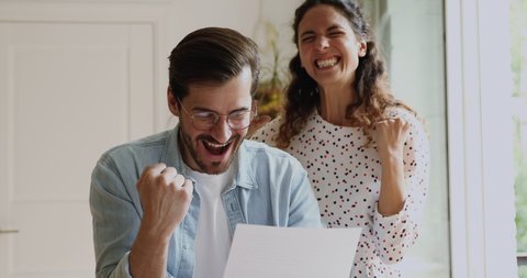 Euphoric millennial family couple reading paper letter with amazing news, celebrating getting bank loan mortgage approval, receiving profitable job offer or promotion, feeling excited indoors.