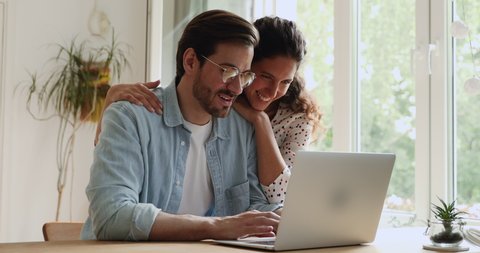 Loving millennial caucasian bonding couple using computer, planning honeymoon or summer vacation, watching family photos, web surfing information or enjoying online shopping together at home.