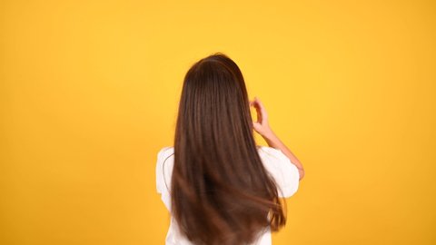 Yong woman shows off her long, straight dark hair on yellow background. Woman stands with her back and touches her hair, healthy even cut. Hair care for health and beauty.