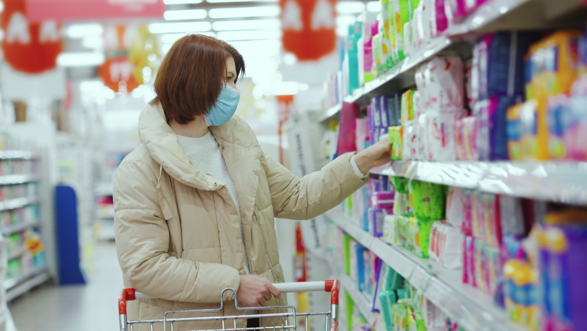 Woman in mask choosing menstrual pads in supermarket, blurred background. Side view young female buying sanitary goods during pandemic. Concept of hygiene | Shutterstock HD Video #1064209594