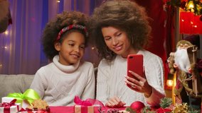 African American mom and little daughter in white sweaters talking via video call on their smartphone. Happy family portrait, New Years celebration concept. Close up.