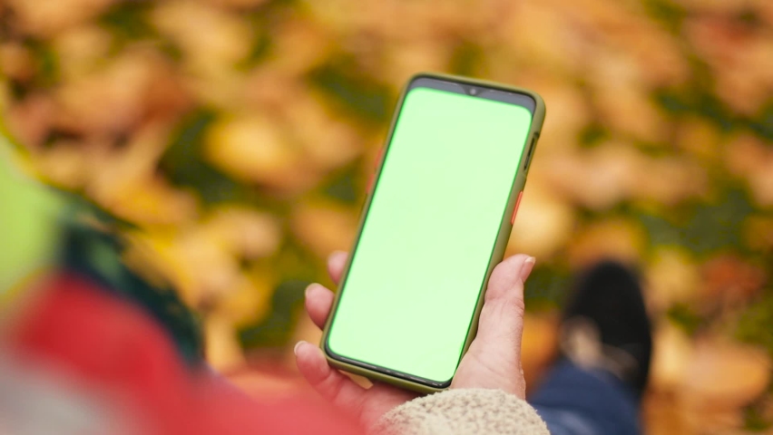 Mock up female hand holding smartphone phone with green, chroma key mobile screen. Girl using mobile phone sitting in autumn park