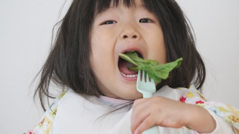 Asian little girl in an apron eating vegetable deliciously by herself. Child eats green lettuce with a plastic fork. Healthy eating and growth of kid. 2 year old toddler baby infant. Isolated on white