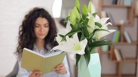 Vase with bouquet of beautiful lilies in room of young woman reading book