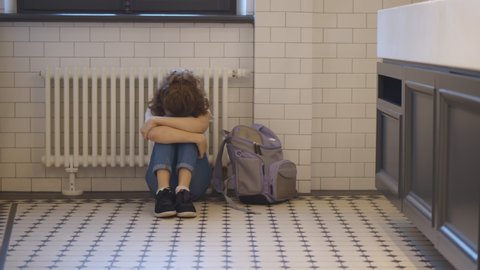 Upset and bullied schoolgirl sitting on floor in restroom and crying. Portrait of depressed preteen girl student in school toilet feeling lonely and sad.