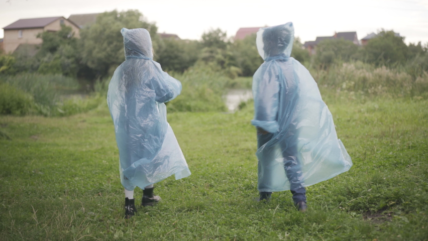 Wide shot of two children in raincoats walking along green summer meadow. Back view of joyful friendly Caucasian friends strolling and talking outdoors. Childhood and friendship.