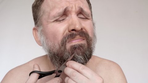 Mature man cuts his beard with scissors and suffers. He is about bust into tears.