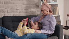 On the lap of her mother with a turban, the little girl is lying on the sofa and making a video call on the phone. Housework, freelance work concept.