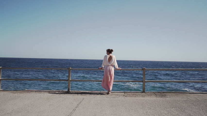 Woman stands next to the blue ocean and admires the beauty of nature. The wind blows the dress. Hot weather. Travel and relaxation on vacation.