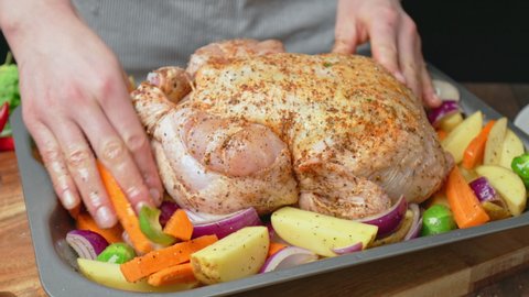 A chef is placing a whole raw marinated chicken into an oven pan with vegetables. Concept of preparing an organic chicken for being grilled.