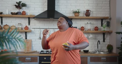 Cheerful plump African-American young woman in orange t-shirt taps on phone screen and starts dancing and singing actively 库存视频