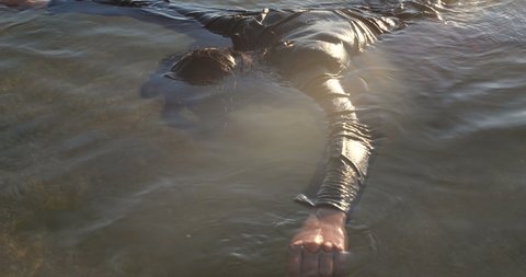 Drowned man with long dark hair in the water sways in the muddy water lying face down in shallow water. Unrecognizable body in black clothes is washed ashore after a shipwreck. 50 fps 4k slow motion