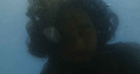 Close-up underwater shot of a long-haired man in black clothes lying in the water and drowned surrounded by jellyfish. The concept of lost hope and suicide. 4k slow motion