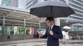 Confidence Asian businessman office worker in suit holding black umbrella walking in the rain with using smartphone. Handsome man business people working in city office district in bad weather day.