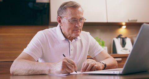 An older man writes his notes onto paper from his laptop. Grandfather learning payment system in internet.