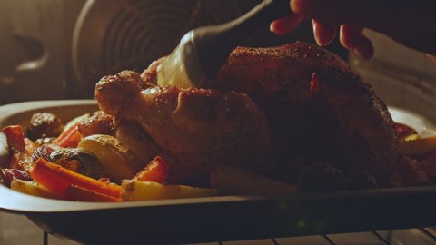 Close-up of brushing and marinating with sauce a whole chicken during grilling. Concept of brushing with a brush a whole organic corn feed chicken along with vegetables into an oven in 4K.