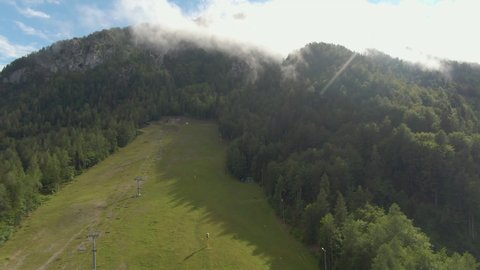 AERIAL, LENS FLARE: Flying over empty ski slopes of Kranjska Gora as mist rises from forest. Cinematic drone view of the misty woods surrounding a ski resort chairlift and slope on a sunny summer day.