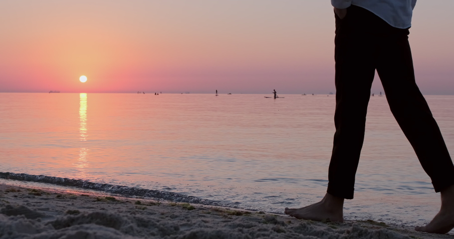 A barefoot man, while walking along the seashore, stops and looks towards the dawn of the sun. A close-up of the legs from a lower perspective. Royalty-Free Stock Footage #1064276164
