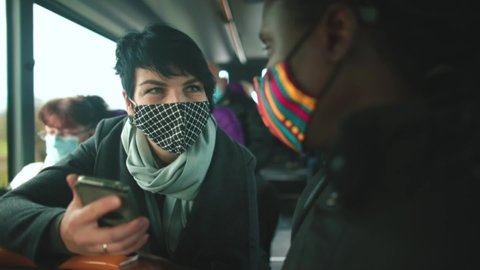 A woman and a black man wearing masks talk on a public transport bus. She looks at phone screen, he tells story of photographs. COVID-19 communication of passengers during transportation.