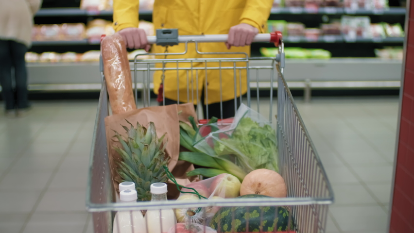 Midsection footage of unrecognizable man in bright yellow coat carrying shopping cart with healthy food products walking along hypermarket