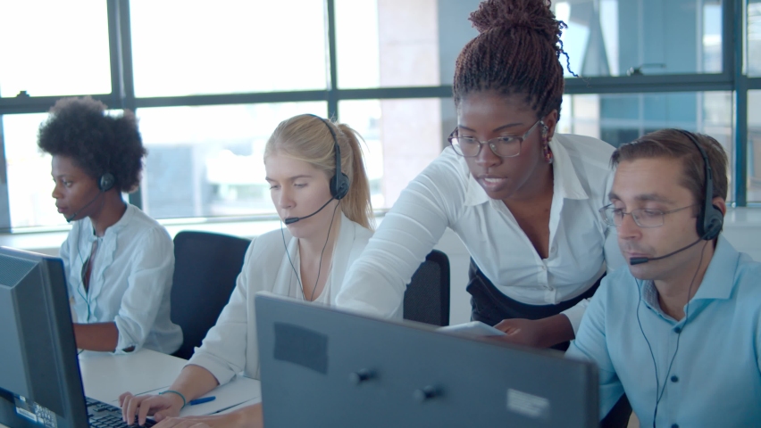 Young African American manager helping call center employees with new software, pointing at monitors and speaking. Customers assistance or telemarketing concept. Royalty-Free Stock Footage #1064299558
