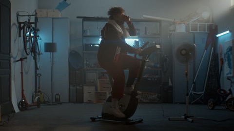 WIDE Funny overweight man in retro outfit drinking water while riding a stationary bicycle in his home garage. Exercising during COVID-19 self isolation quarantine