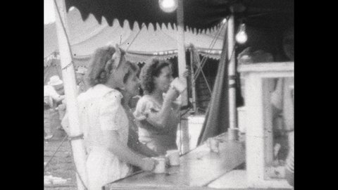 1940s: Women idle at circus concession stands enjoying treats; man stands in circus ticket booth.