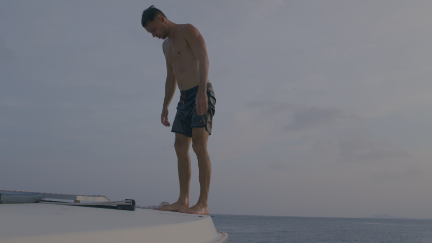 Young man jumping off cliff into blue water ocean at sunset. Active outdoor, holiday adventure, tourism action, healthy summer joy, Fun activity lifestyle. Crazy adult guy in swimwear fly from climb