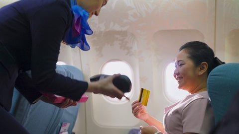 Onboard product purchasing, Asian woman sitting in airplane buying gift from air hostess and paying by credit card tapping on machine. Contactless payment and technology inflight service concept.