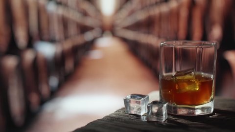 A glass of whiskey with ice is on a wooden table. In the background are barrels of whiskey.