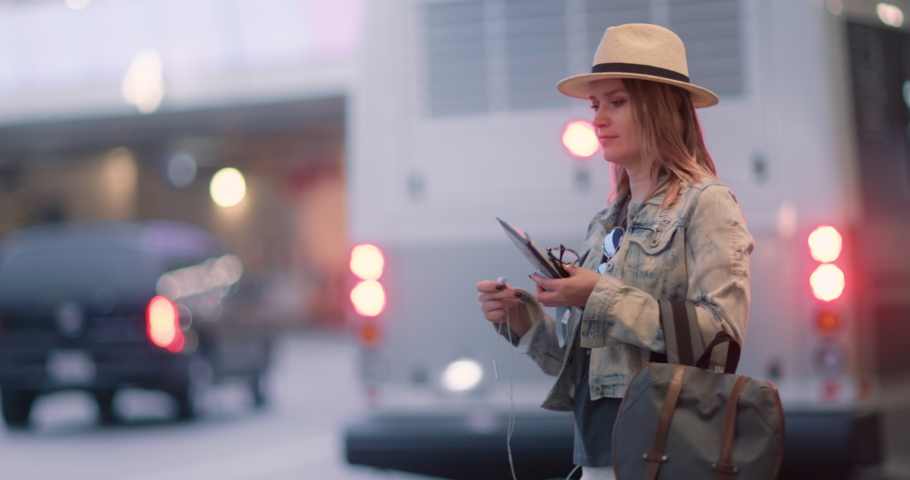 Traveler listen music while waiting the taxi at loading zone at the arrival exit of airport terminal. Slow motion woman with backpack and travel documents enjoying a song via headset, 4K