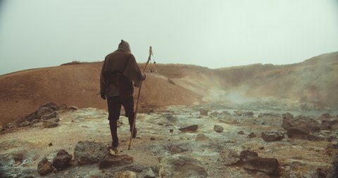 Handheld Slow Motion Wide Shot Of Hooded Man In Robes Carrying Staff Through Mysterious Desert Landscape.