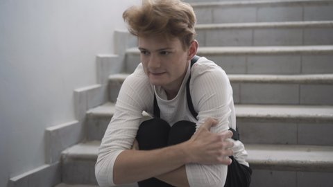 Close-up portrait of scared bullied redhead boy sitting on stairs shaking. Frightened insulted Caucasian high school student hiding from bullies in public school. Mocking and bullying.