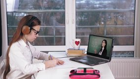 distance learning, online schooling. schoolgirl watching online tutorial video lesson with teacher's explanations on laptop and making notes in copybook. studying online at home.