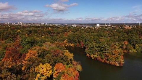 Beautiful aerial flying over the lakes and fall foliage covered trees in Roger Williams Park with the Providence, Rhode Island downtown skyline and river seen beyond.