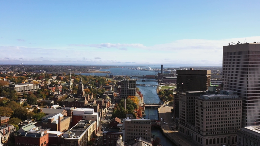Aerial fly over above the high rise buildings following the Moshassuck and Providence Rivers in downtown Providence, Rhode Island with smokestacks, bridges, wind turbines and church steeples beyond.