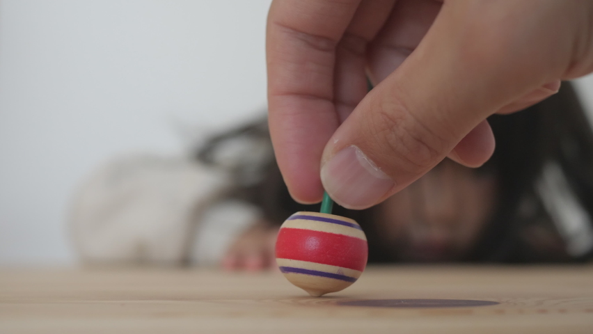 A small wooden spinning top colored red and blue on a board in the background of Asian little girl. Parent and toddler playing together at home. Funny kid tries to stop the top with index finger
