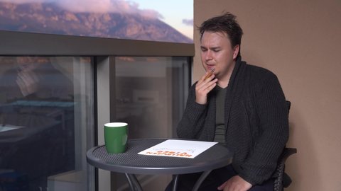 Young male with tousled hair wearing casual, sits in front of the window with beautiful mountain view and try to create Halloween concept on the sheet of paper. Creative crisis or difficulty