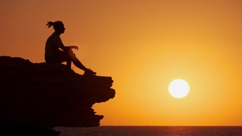 Silhouette of hipster woman puts headphones and enjoys music while sitting on cliff face looking at sea at sunset.