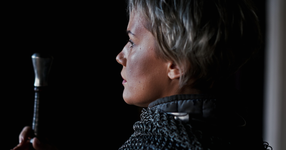 Close up on a woman warrior in chain mail armour with focus to the side of her face against a dark shadowy background