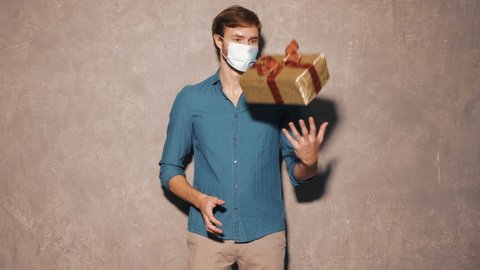 Handsome smiling hipster lumbersexual businessman model wearing casual jeans shirt clothes. Fashion stylish man posing  near gray wall in studio. In protective mask for corona virus. Holding gift box