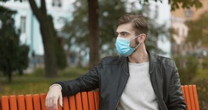 Sick young man sits on the bench in sity park. Then he takes off medical protective mask, he is not feeling well and has a headache, he massages his head. Cinema 4K Slow Motion Video
