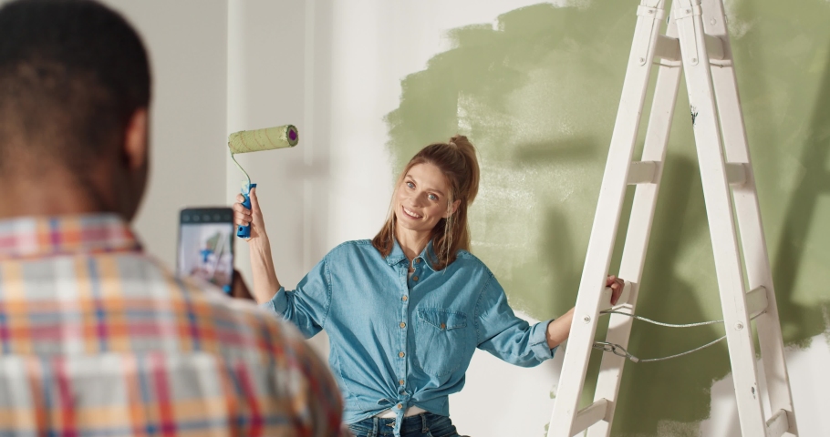Rear of African American husband taking photos of beautiful joyful Caucasian wife on smartphone posing in new home holding brush roller repairing and painting wall. Remodeling and redesigning concept