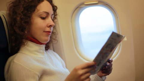 young woman sits in airplane and reading magazine, then looks out window