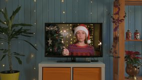 Happy woman with sparklers and santa hat congratulates relatives on Christmas via smart tv online.